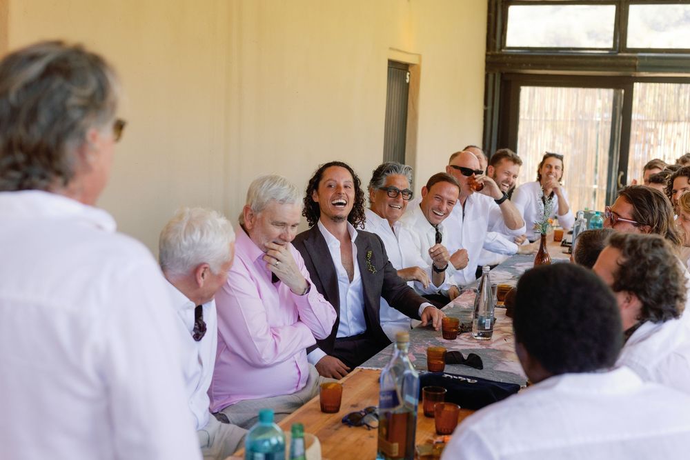 Wedding guests laughing and socializing together at a long table during an outdoor daytime reception.