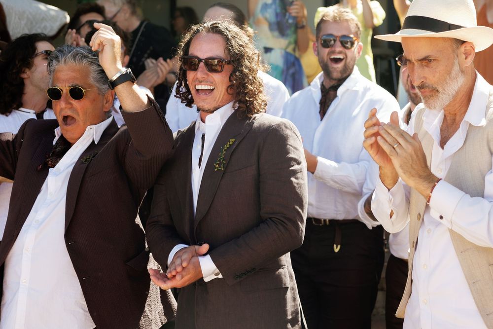Groom in brown suit smiling and celebrating with guests clapping during an outdoor wedding ceremony.
