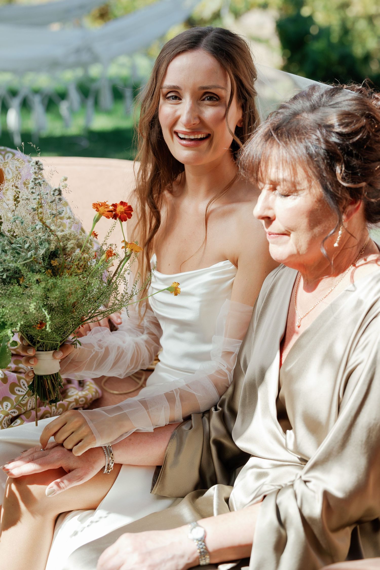 Smiling bride in strapless gown holding wildflower bouquet sits beside older woman in sage dress outdoors.