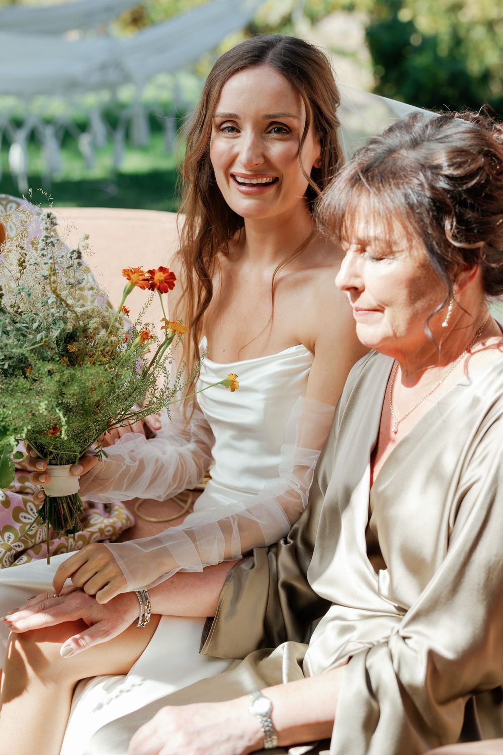 Smiling bride in strapless gown holding wildflower bouquet sits beside older woman in sage dress outdoors.