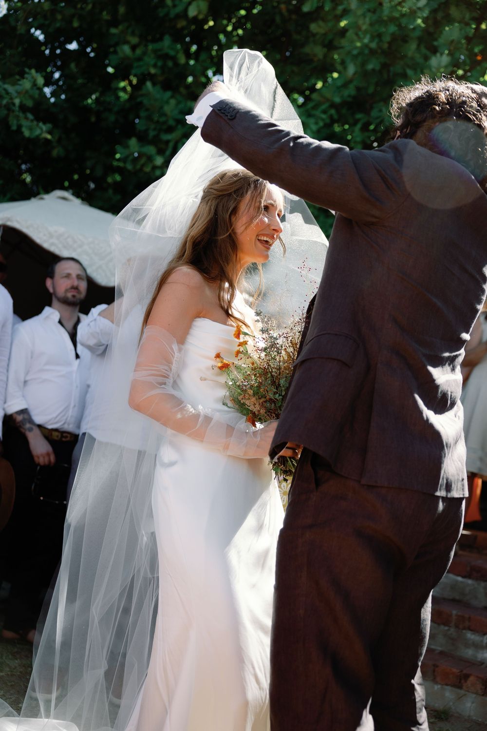 Groom lifting veil over laughing bride in white strapless dress during sunny outdoor wedding ceremony.
