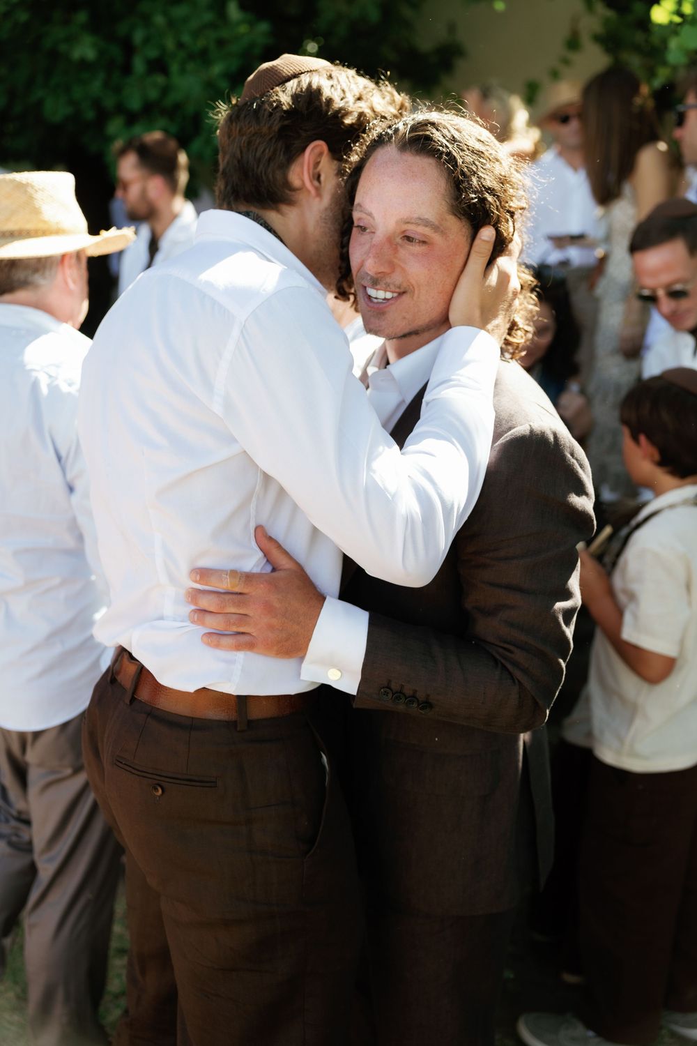 Two men in brown trousers embracing warmly at an outdoor gathering with guests in background.