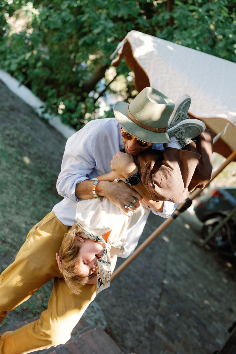 Aerial view of couple embracing outdoors, woman in yellow pants and man in hat, sunny day.