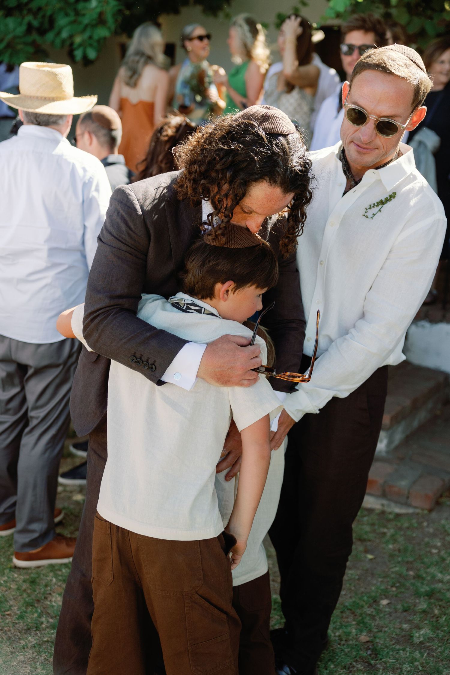 Man in brown suit hugging child at outdoor wedding reception while guest in white shirt looks on.