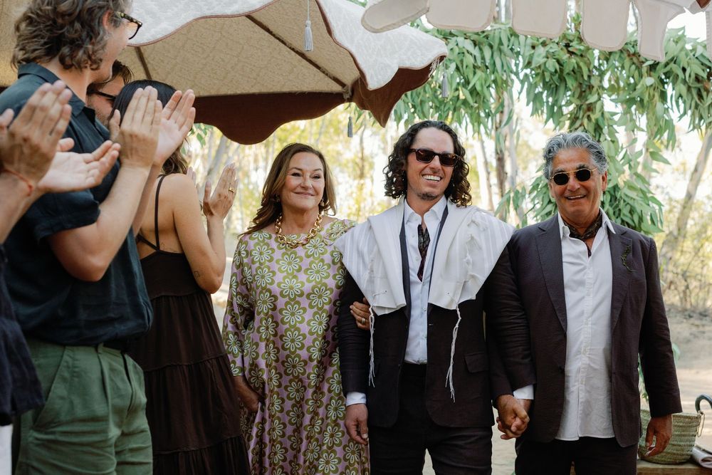 Groom wearing tallit standing with family members under a chuppah during a Jewish wedding ceremony.