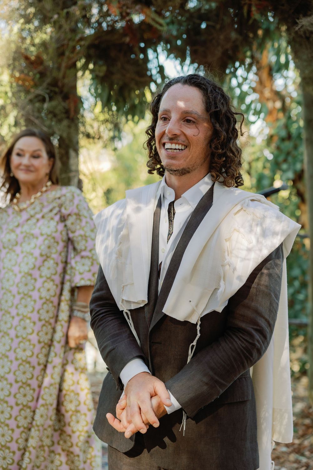 Smiling groom in dark suit and white tallit laughs joyfully during outdoor Jewish wedding ceremony.