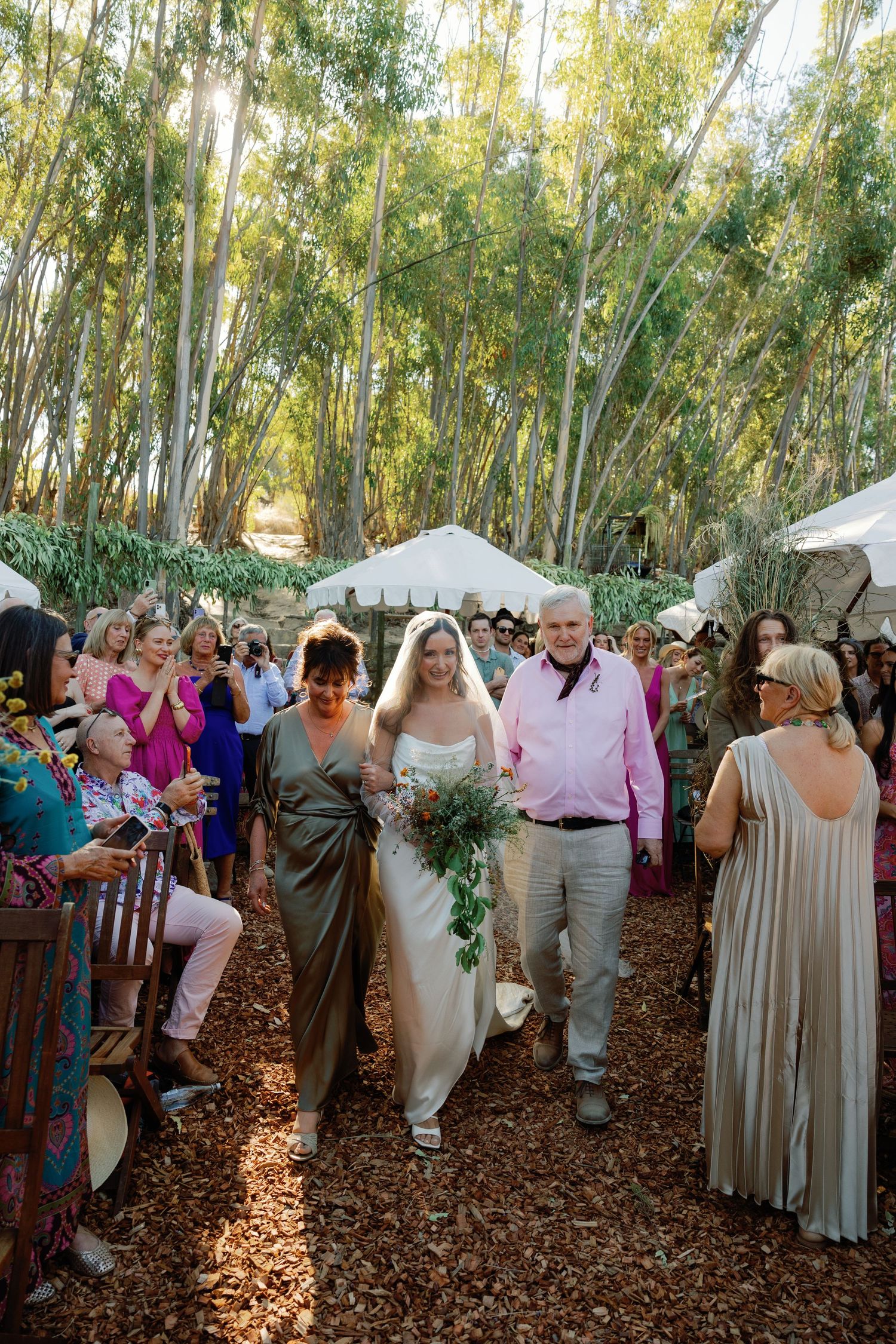 Bride in white gown walks down wooded aisle with parents at outdoor wedding ceremony.