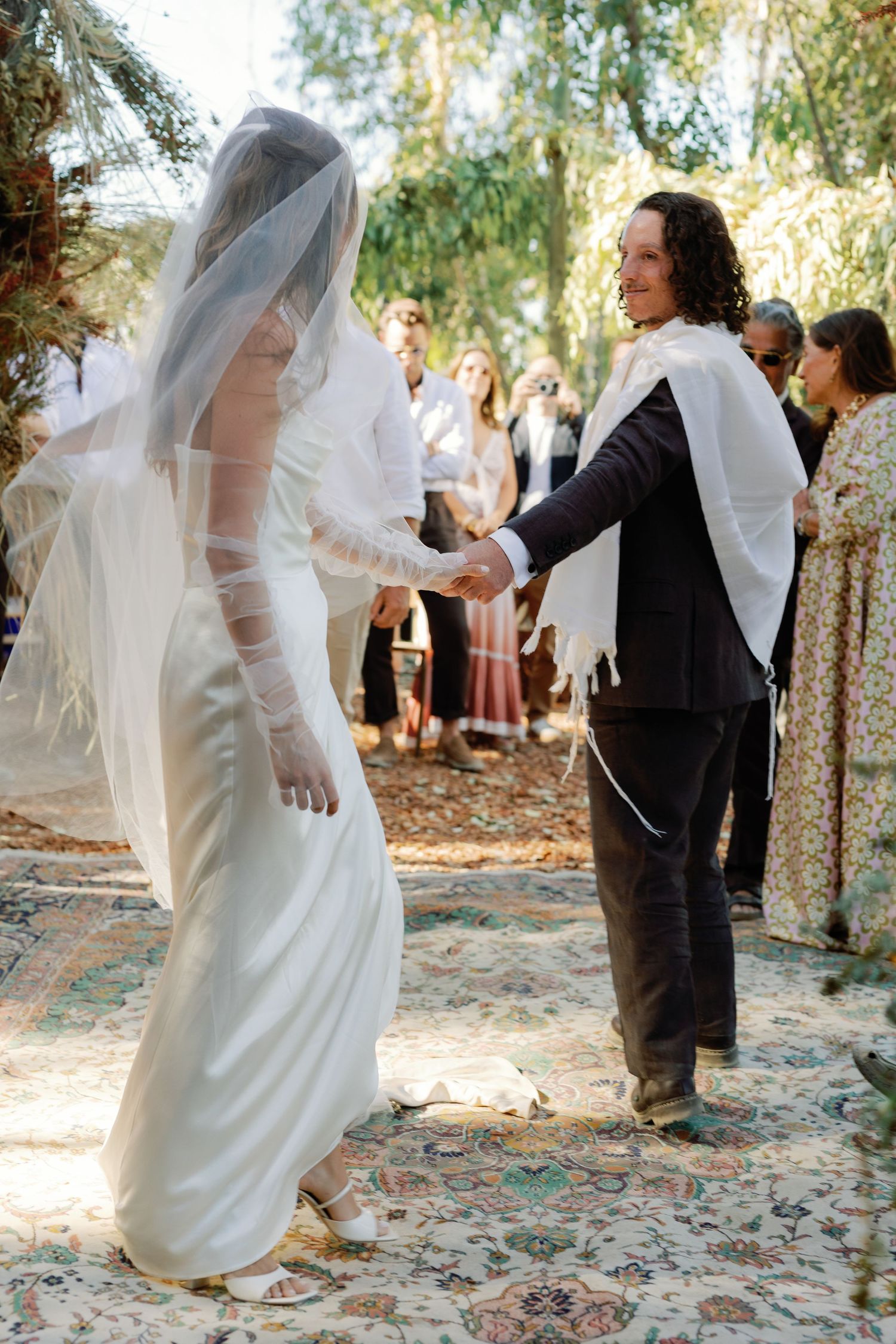 Bride and groom hold hands during an outdoor wedding ceremony surrounded by guests under trees.