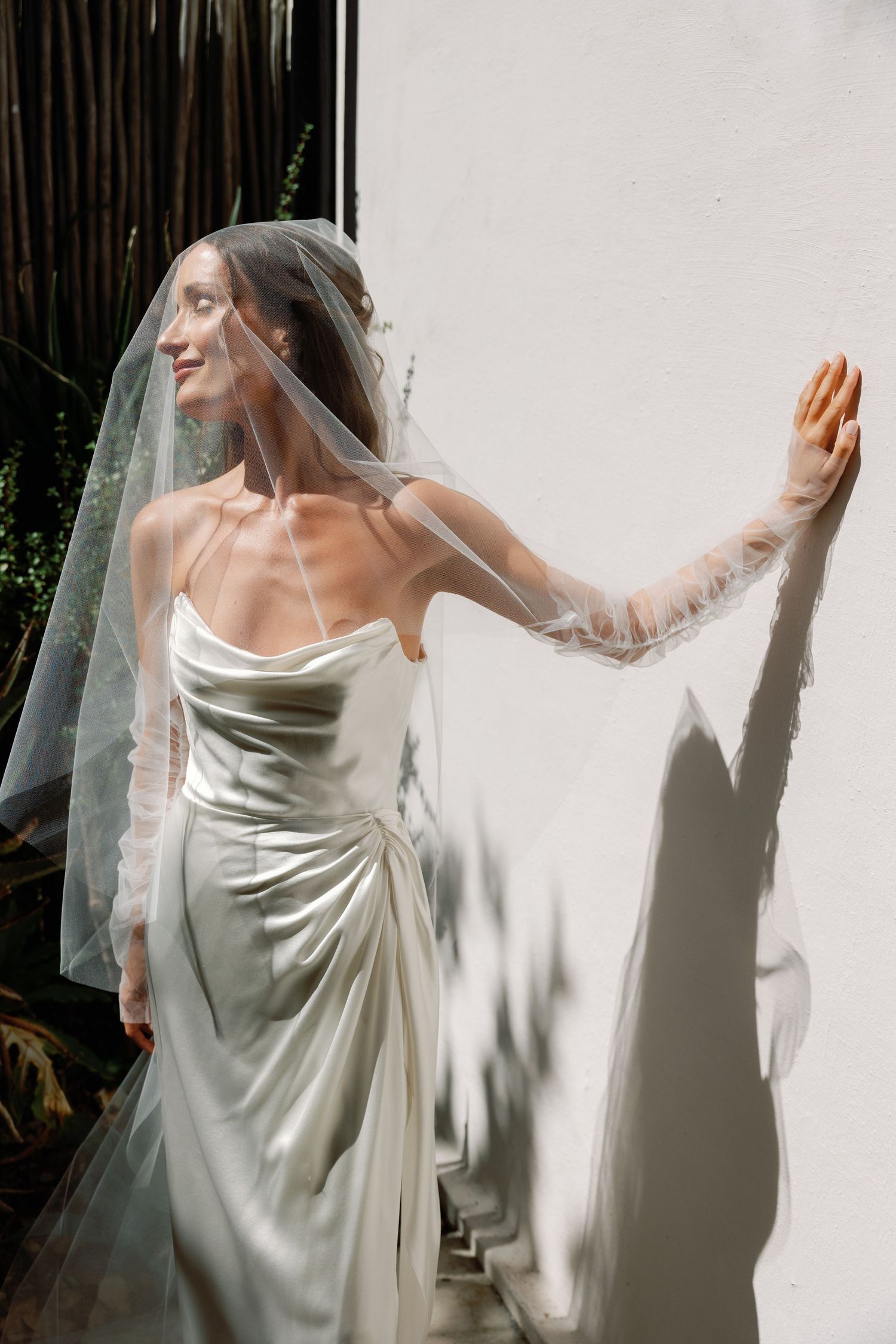 Bride in satin slip gown and sheer veil leans against white wall in sunlight, eyes closed, smiling softly.