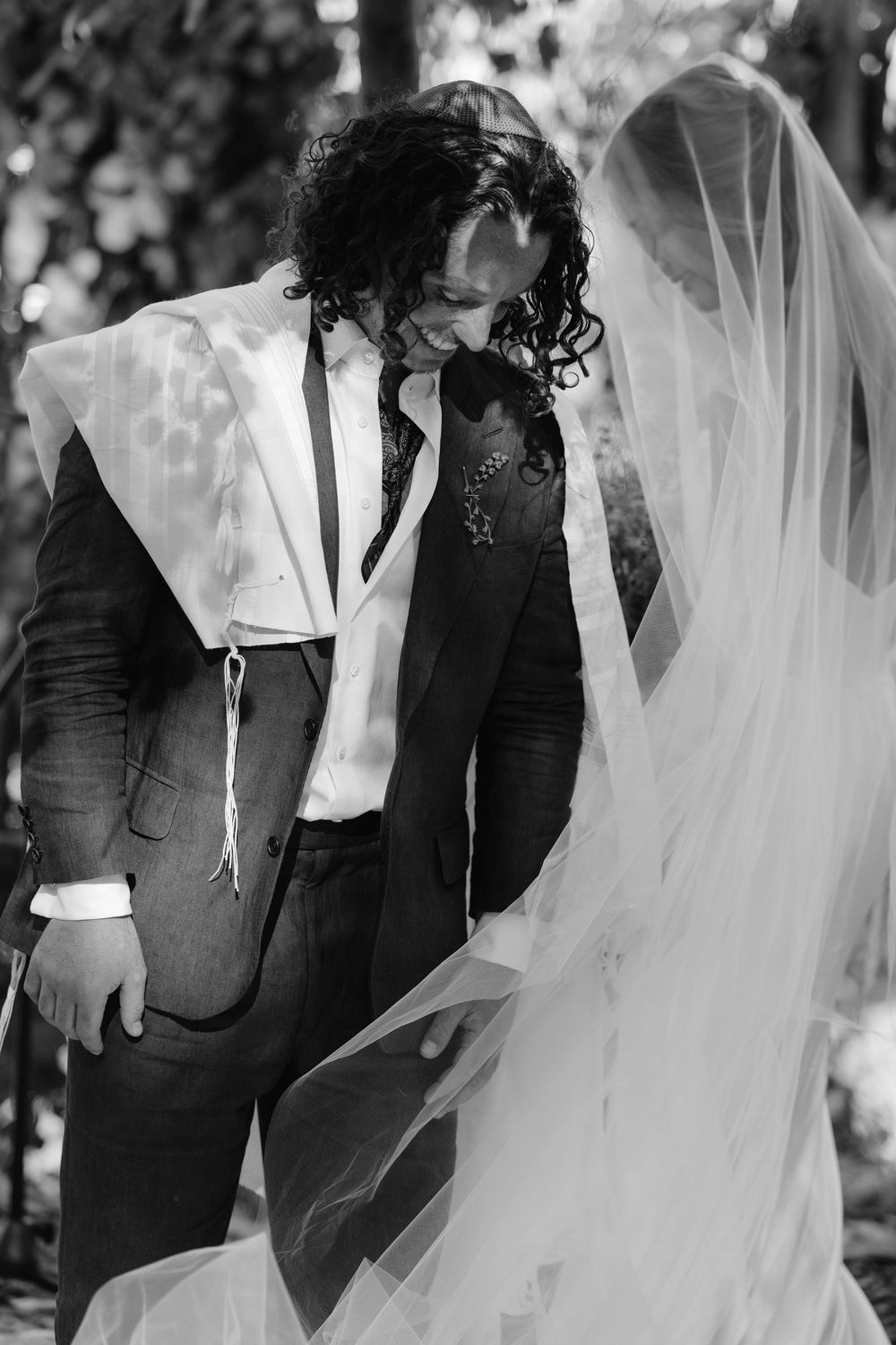 Groom with curly hair smiling at veiled bride during outdoor wedding ceremony, black and white.