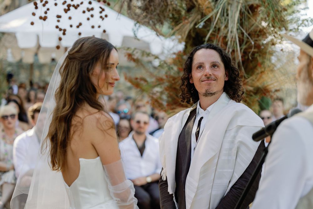Groom in white tallit smiling at bride during outdoor Jewish wedding ceremony under chuppah.