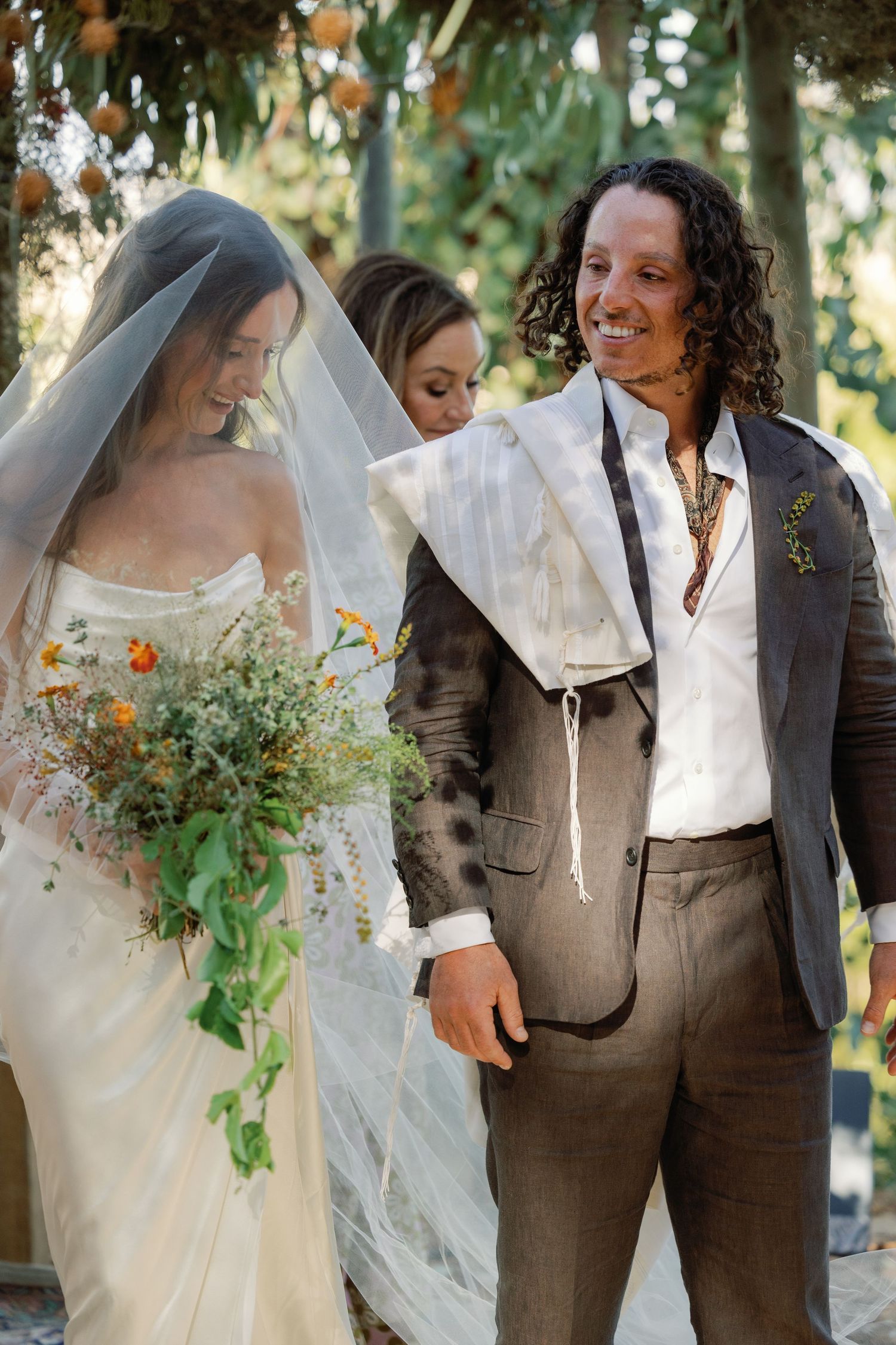 Smiling groom with curly hair and bride in veil holding wildflower bouquet at outdoor ceremony.