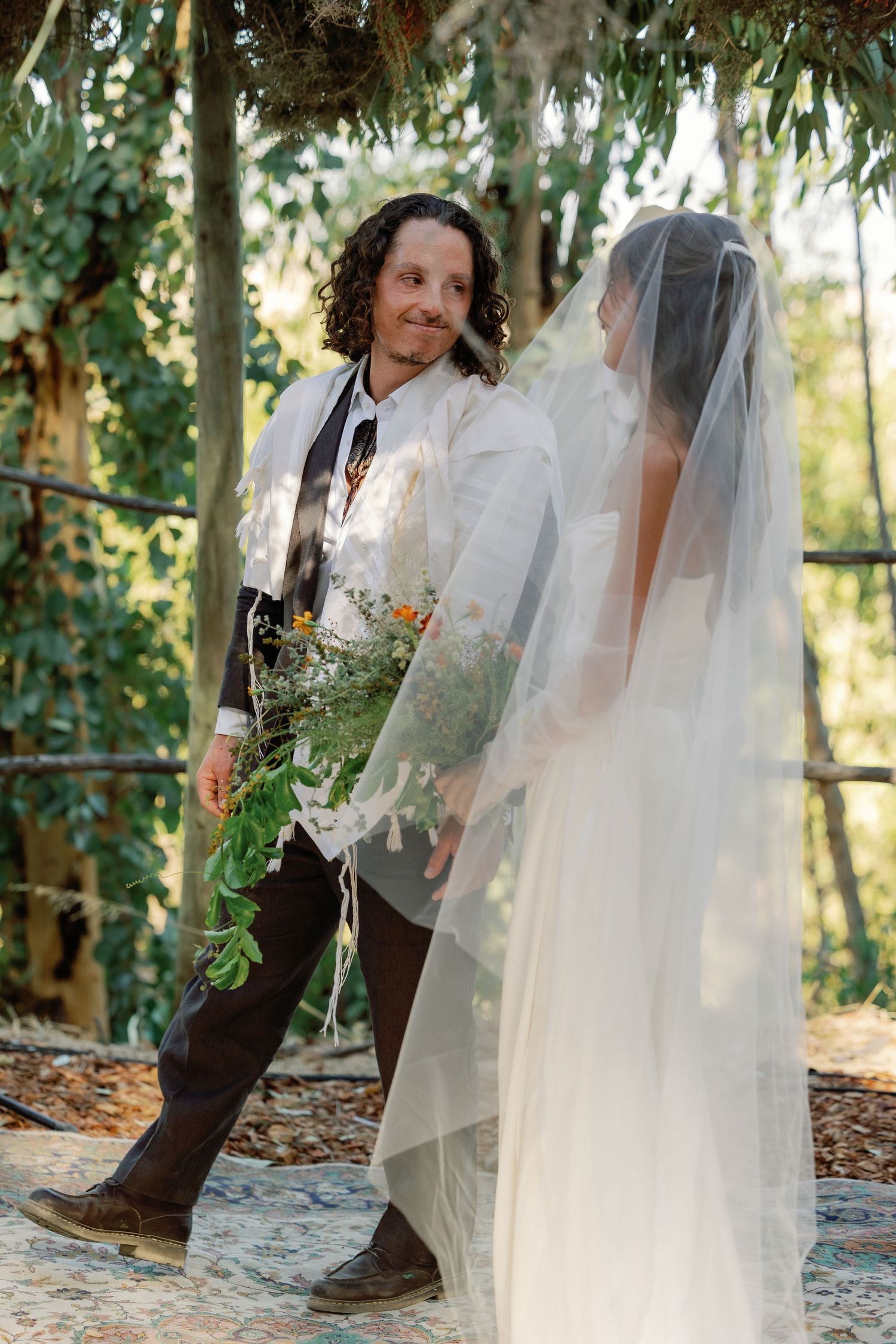 Groom in tallit holds wildflower bouquet facing veiled bride during outdoor Jewish wedding ceremony.