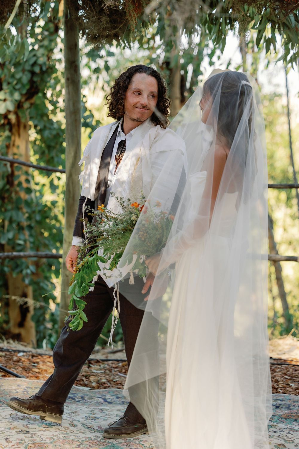 Groom in tallit holds wildflower bouquet facing veiled bride during outdoor Jewish wedding ceremony.