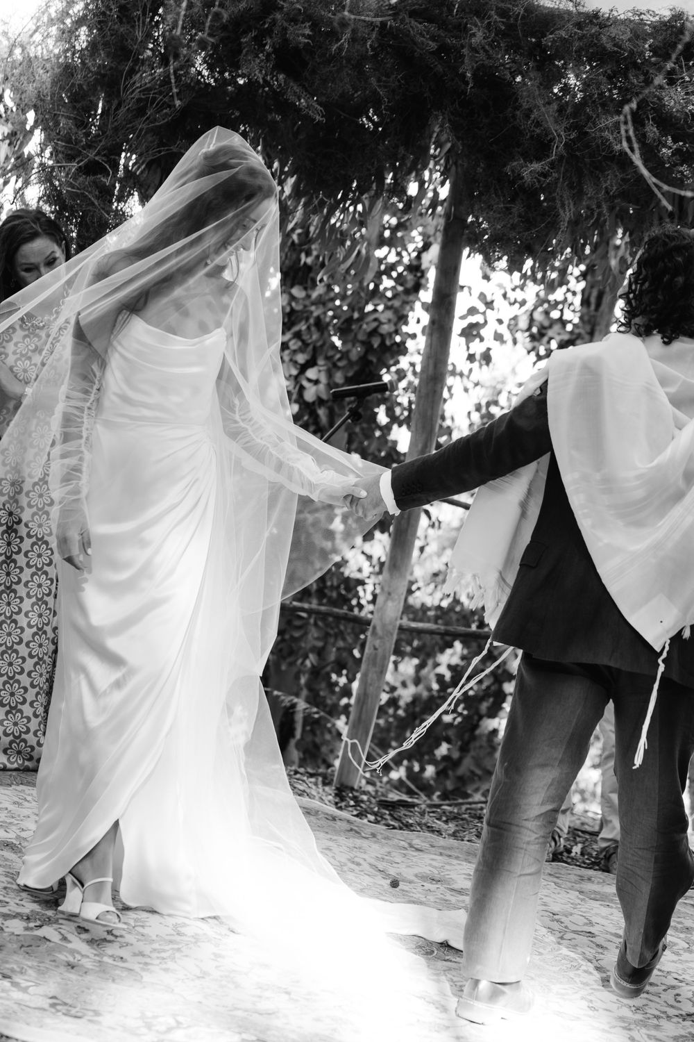 Bride in white gown and veil holding hands with groom during outdoor wedding ceremony, black and white.