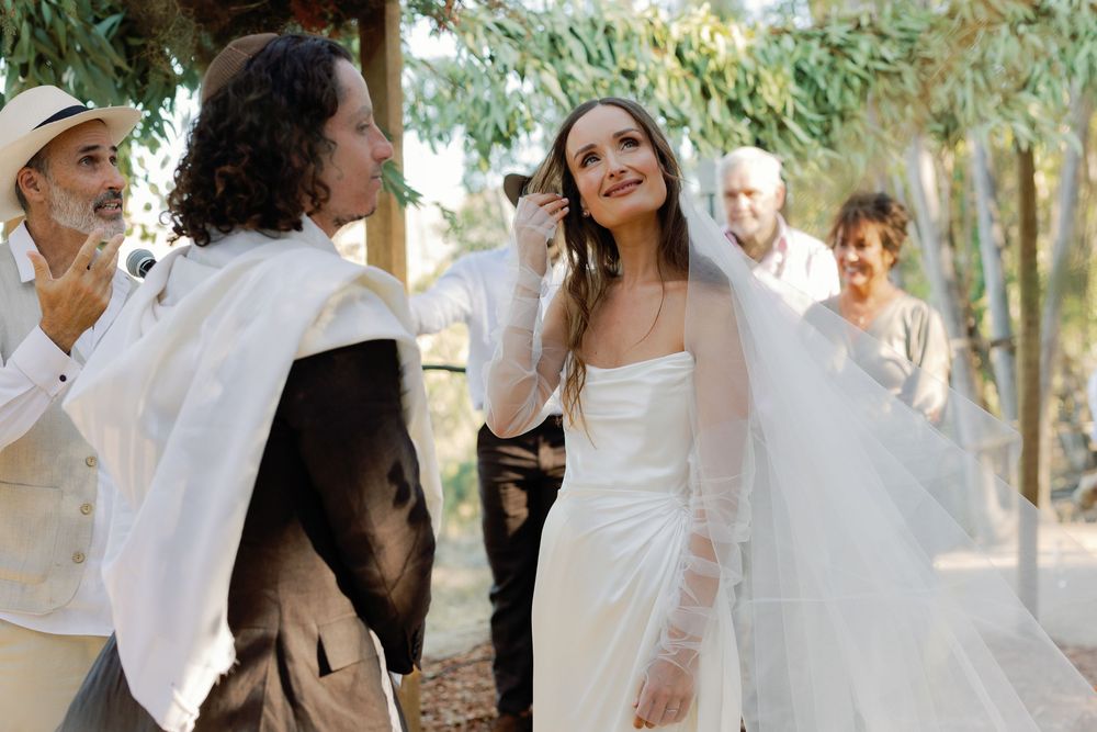 A smiling bride in a white gown and veil stands with guests during an outdoor wedding celebration.