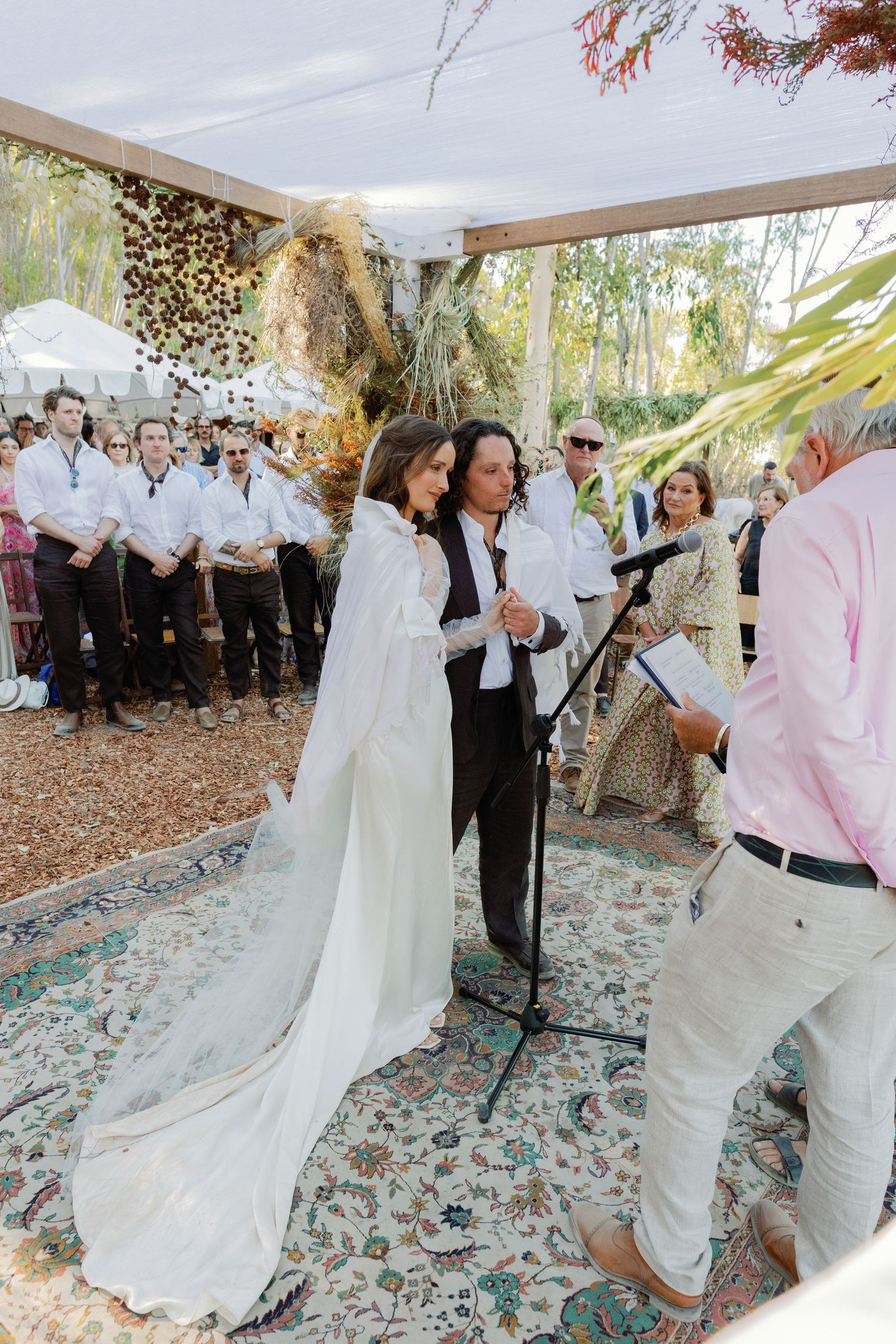 A bride in a white cape gown stands with her partner at an outdoor wedding ceremony under a canopy.