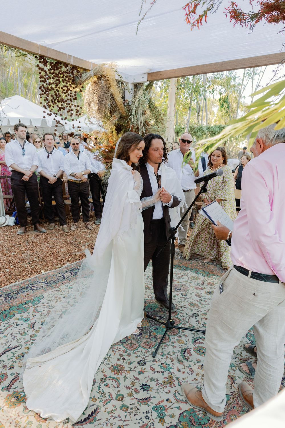 A bride in a white cape gown stands with her partner at an outdoor wedding ceremony under a canopy.
