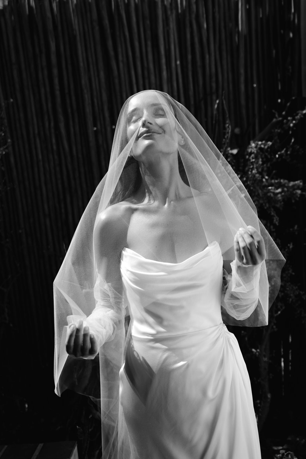 Black and white photo of a bride in a white gown and veil, head tilted back with eyes closed outdoors.