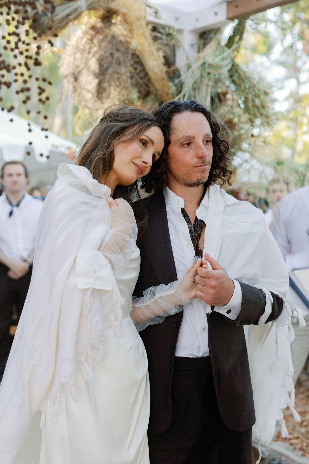 Bride and groom share intimate moment wrapped together in white tallit at outdoor Jewish wedding.