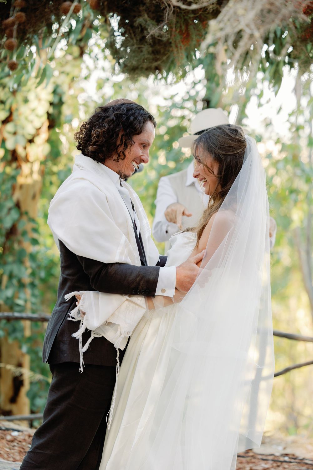 Bride and groom laughing together under floral chuppah at outdoor Jewish wedding ceremony.