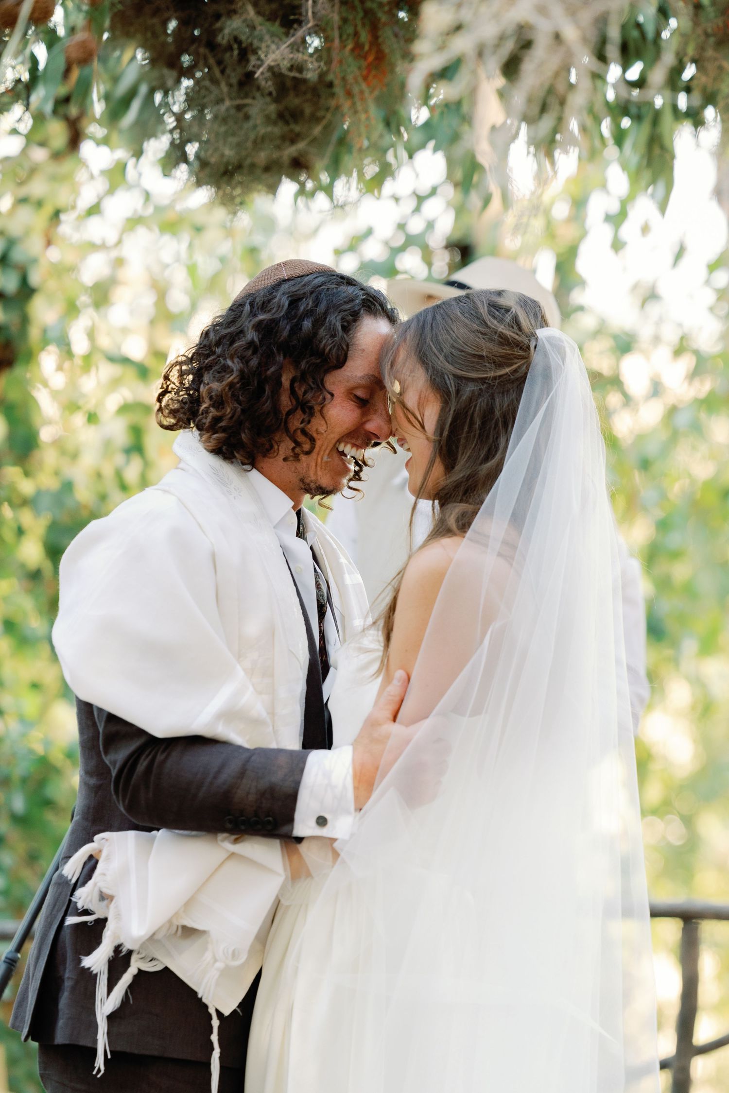 Newlyweds share intimate forehead-touching moment under wedding canopy with lush green foliage backdrop.