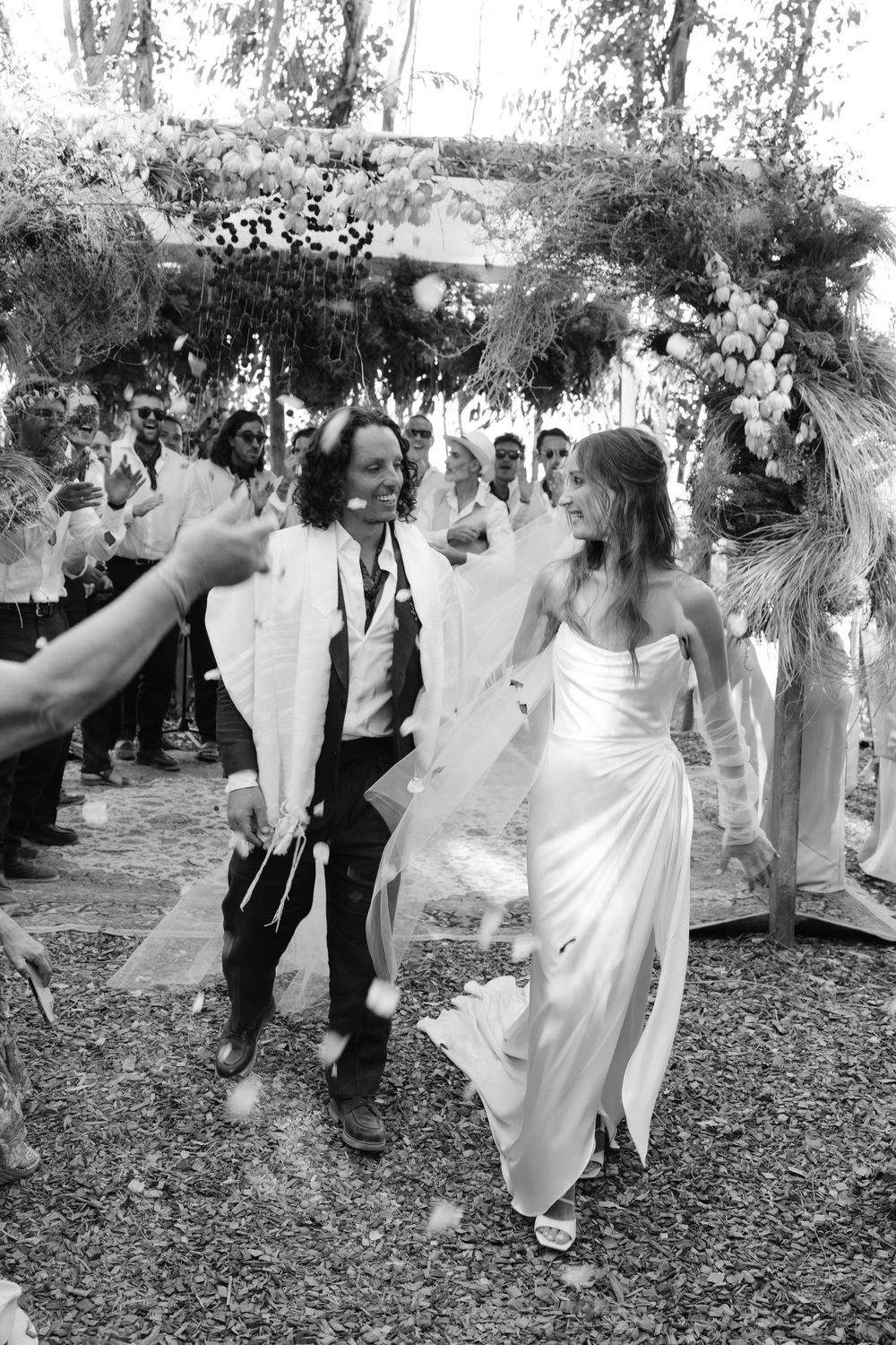 Black and white photo of newlyweds celebrating as they exit wedding ceremony under floral arch.