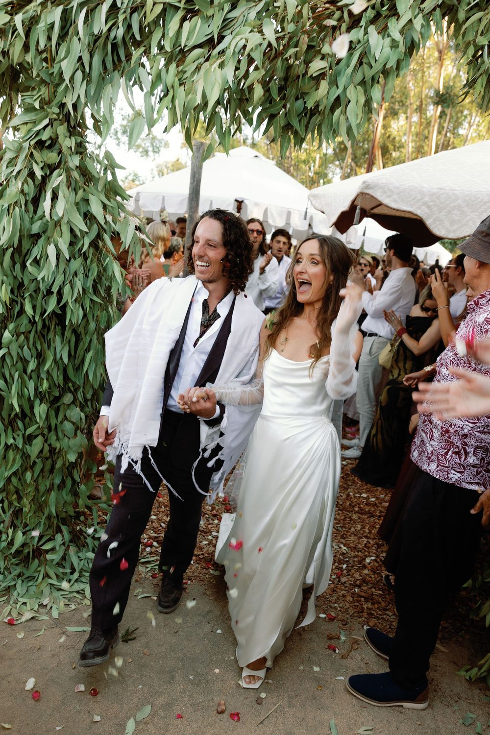 Joyful newlyweds walk through petal shower under eucalyptus arch surrounded by cheering wedding guests.