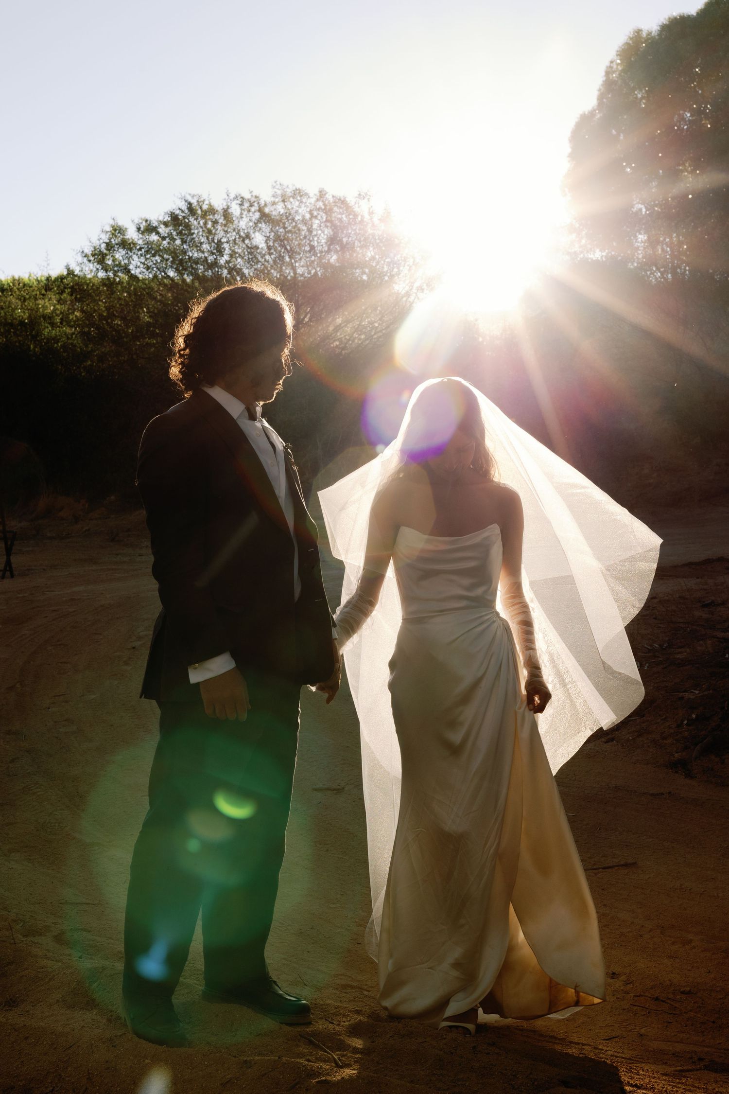 Bride and groom holding hands walking into golden sunset backlight at outdoor wedding venue.