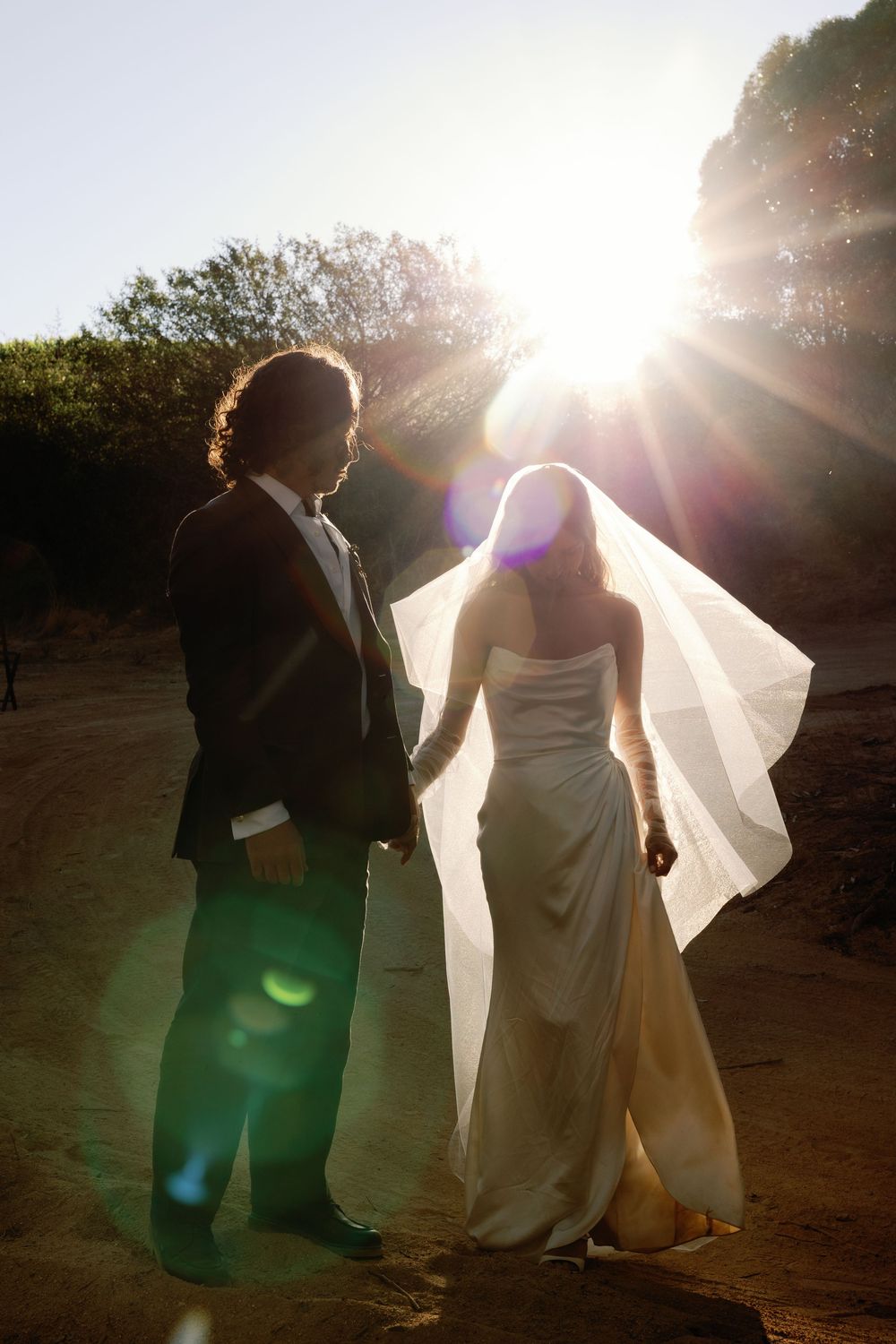 Bride and groom holding hands walking into golden sunset backlight at outdoor wedding venue.