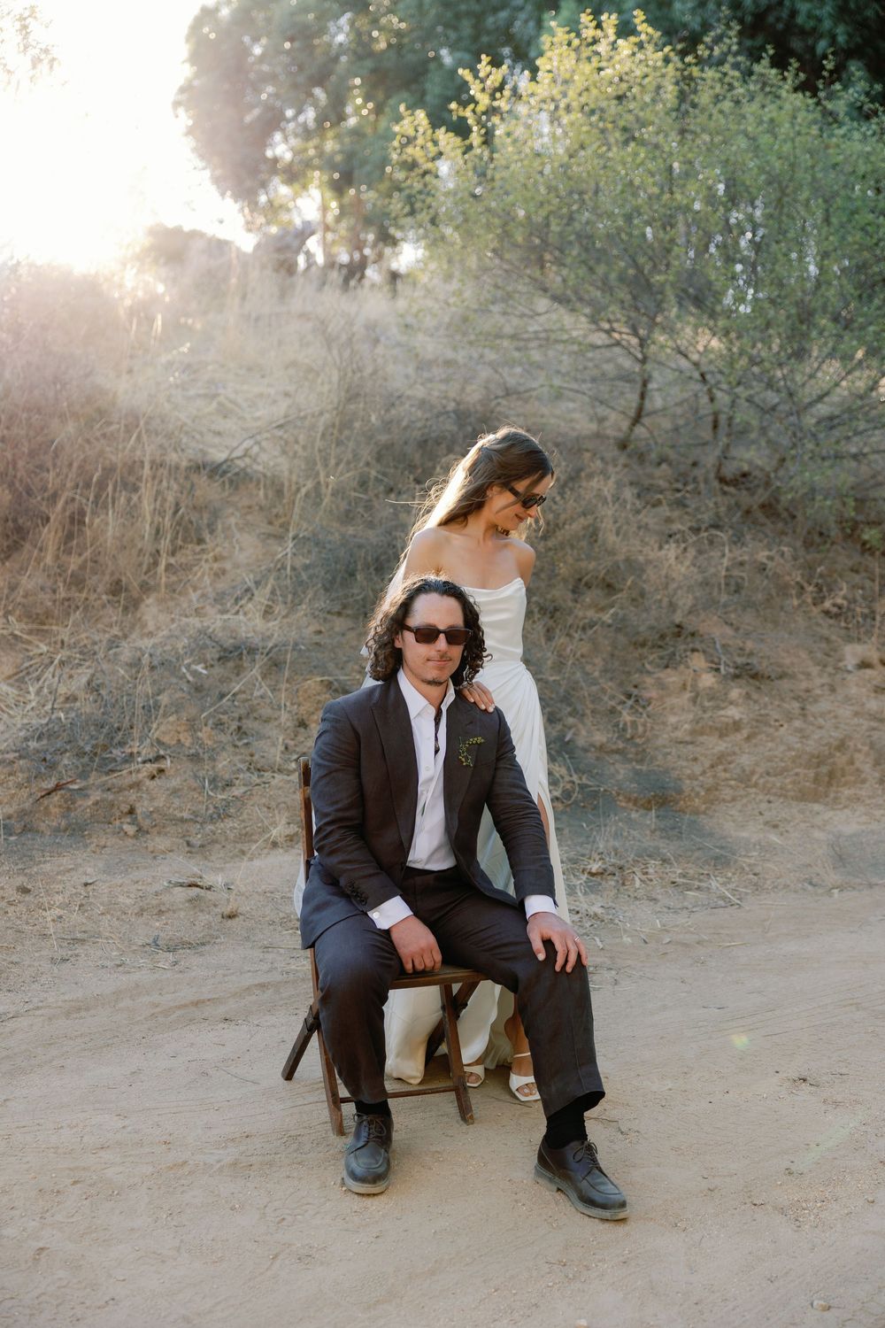 A groom in a dark suit sits on a chair while the bride stands behind him in a sunlit outdoor setting.