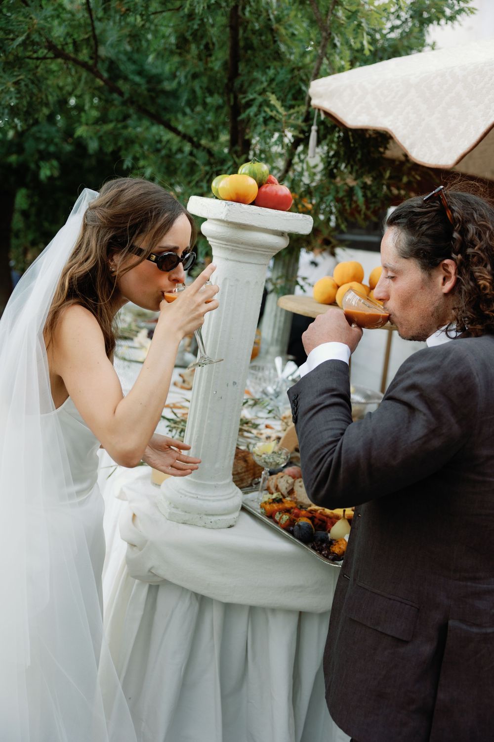 Bride in sunglasses and groom in suit eating at an elegant outdoor wedding reception buffet table.