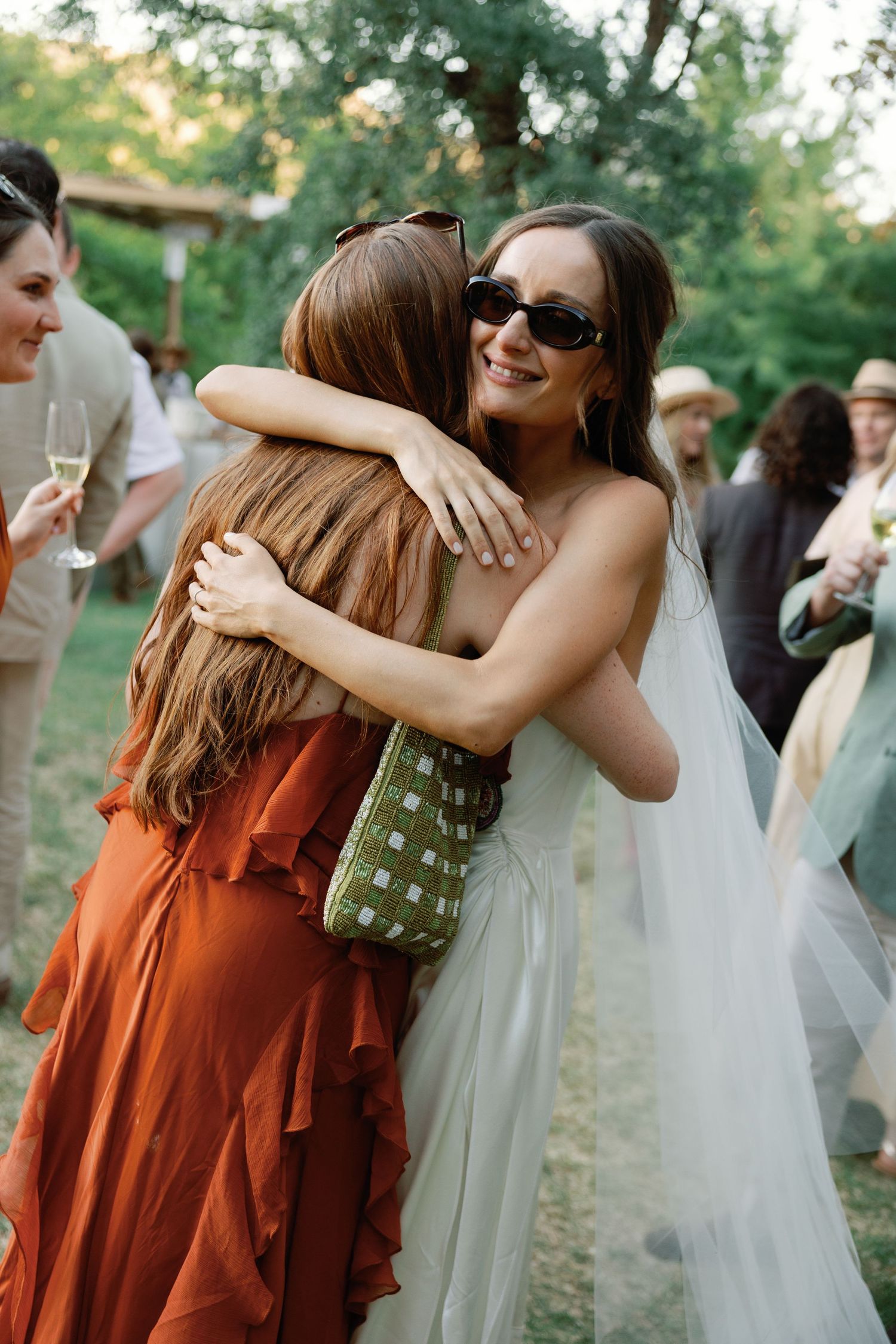 Bride in white gown and veil hugging female guest in orange dress at outdoor wedding reception.
