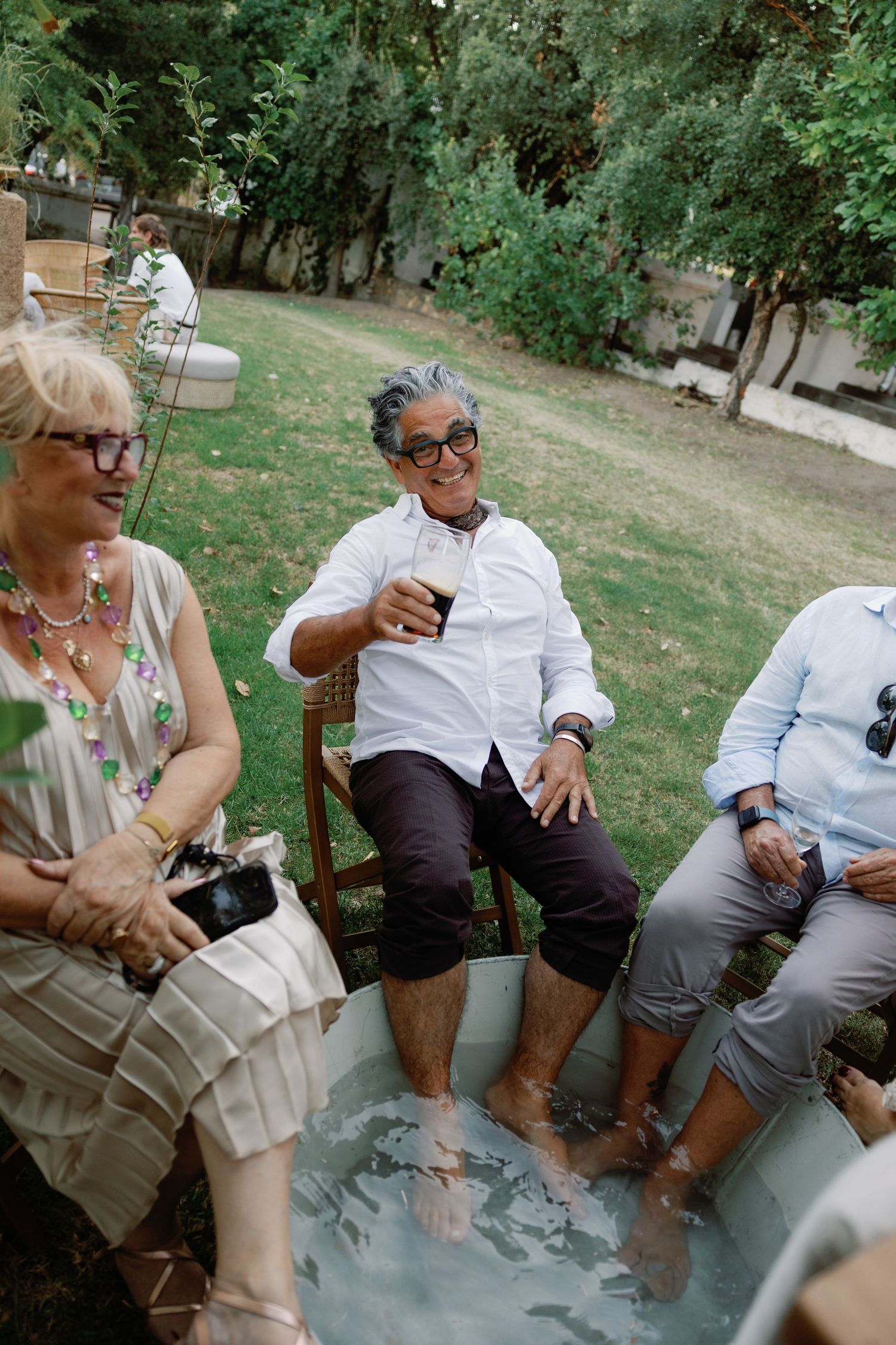 Wedding guests enjoy a foot bath in a garden setting while socializing and drinking at an outdoor reception.