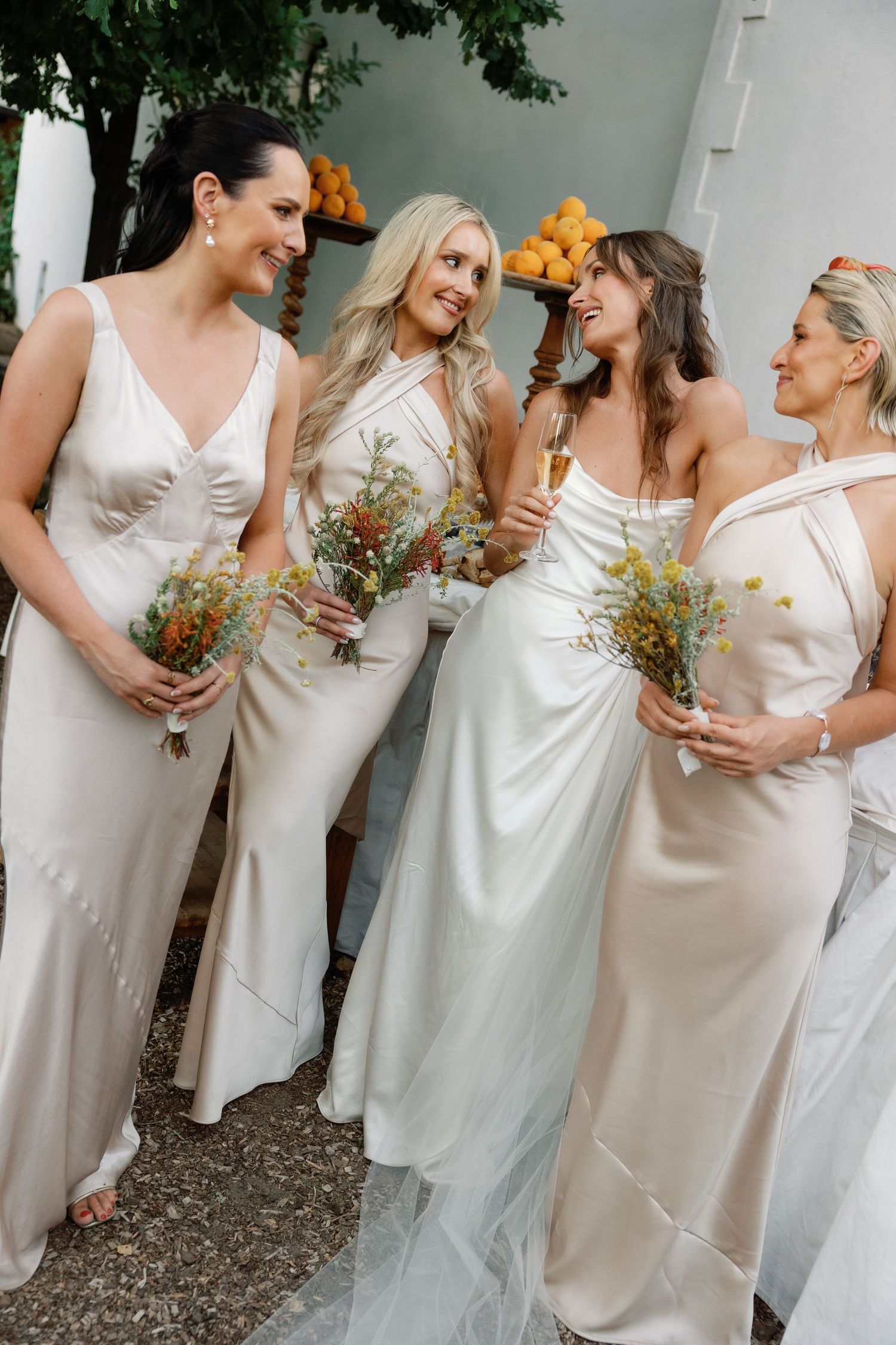 Bride and three bridesmaids in champagne satin gowns holding wildflower bouquets at an outdoor wedding.