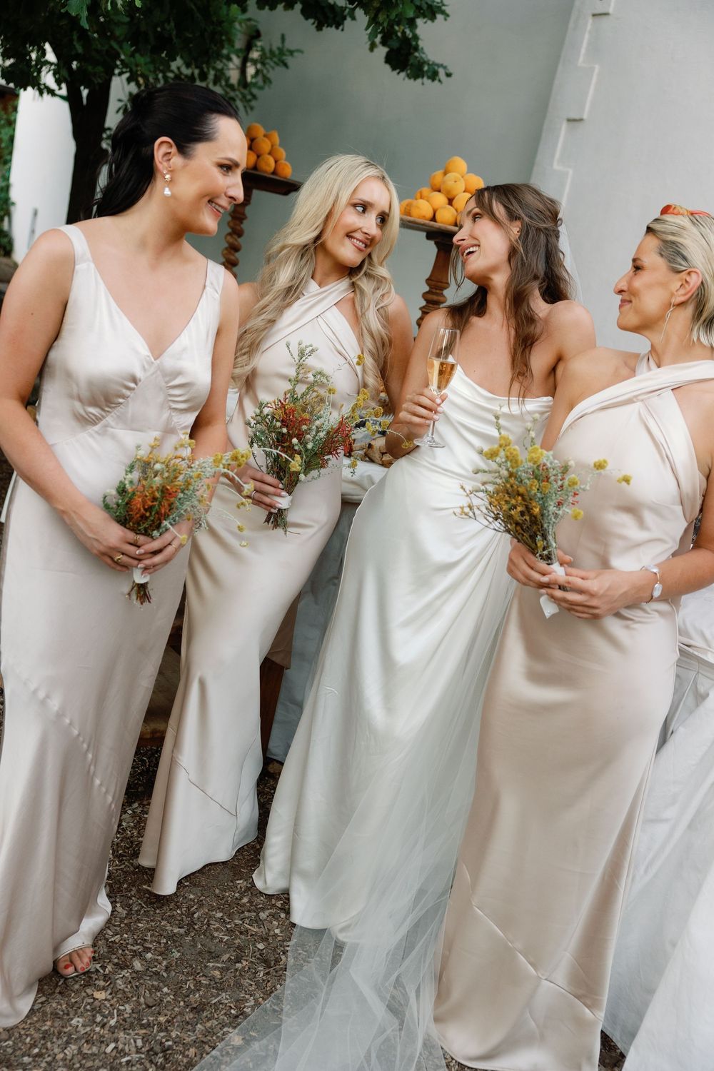 Bride and three bridesmaids in champagne satin gowns holding wildflower bouquets at an outdoor wedding.