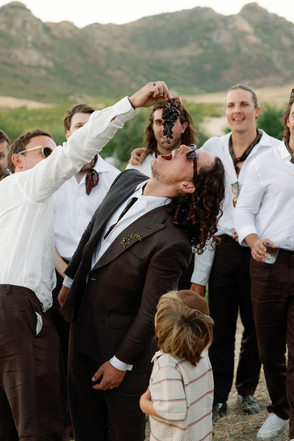 Groom in dark suit eating grapes held above his head, surrounded by groomsmen in white shirts outdoors.