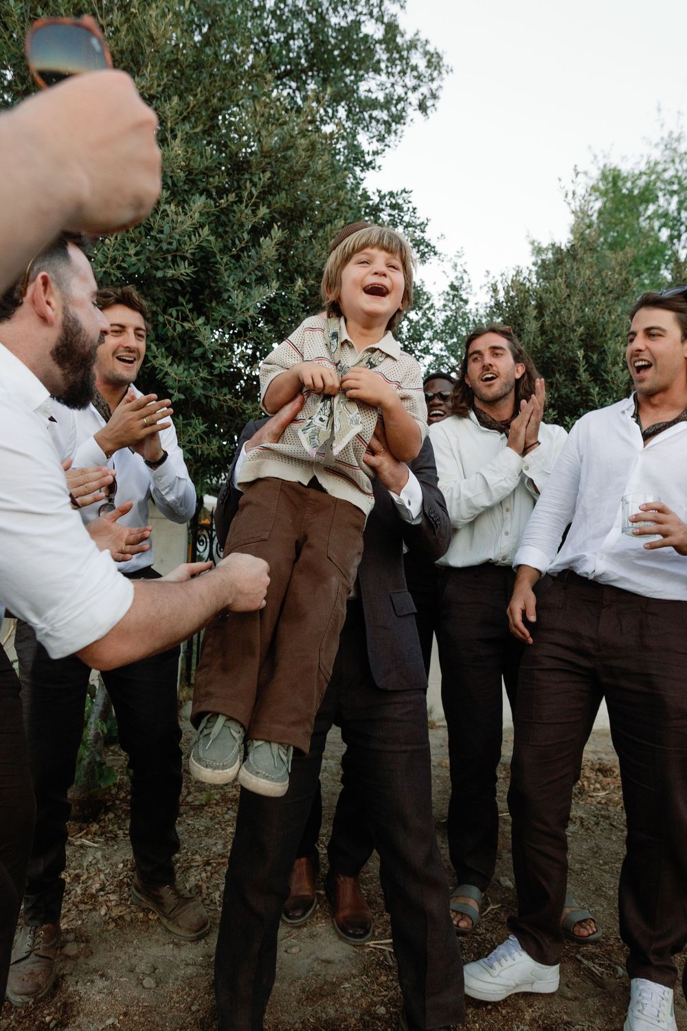 A joyful boy is lifted by laughing groomsmen outdoors at a wedding celebration surrounded by trees.