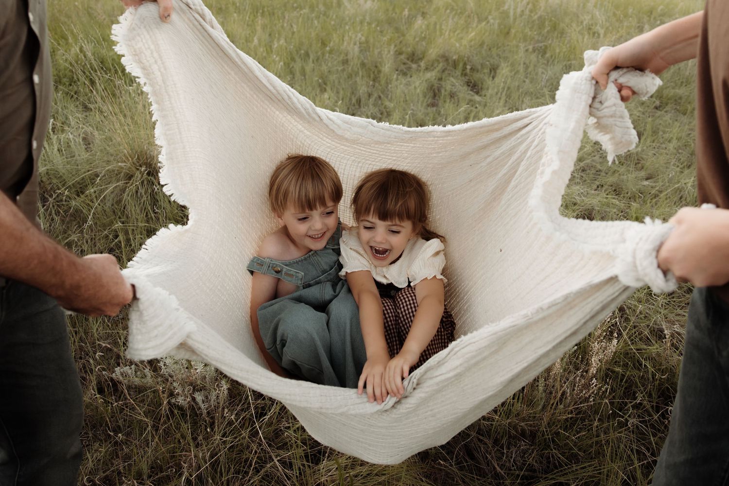 Two young girls laughing together in a white fabric hammock held by adults in a green field.