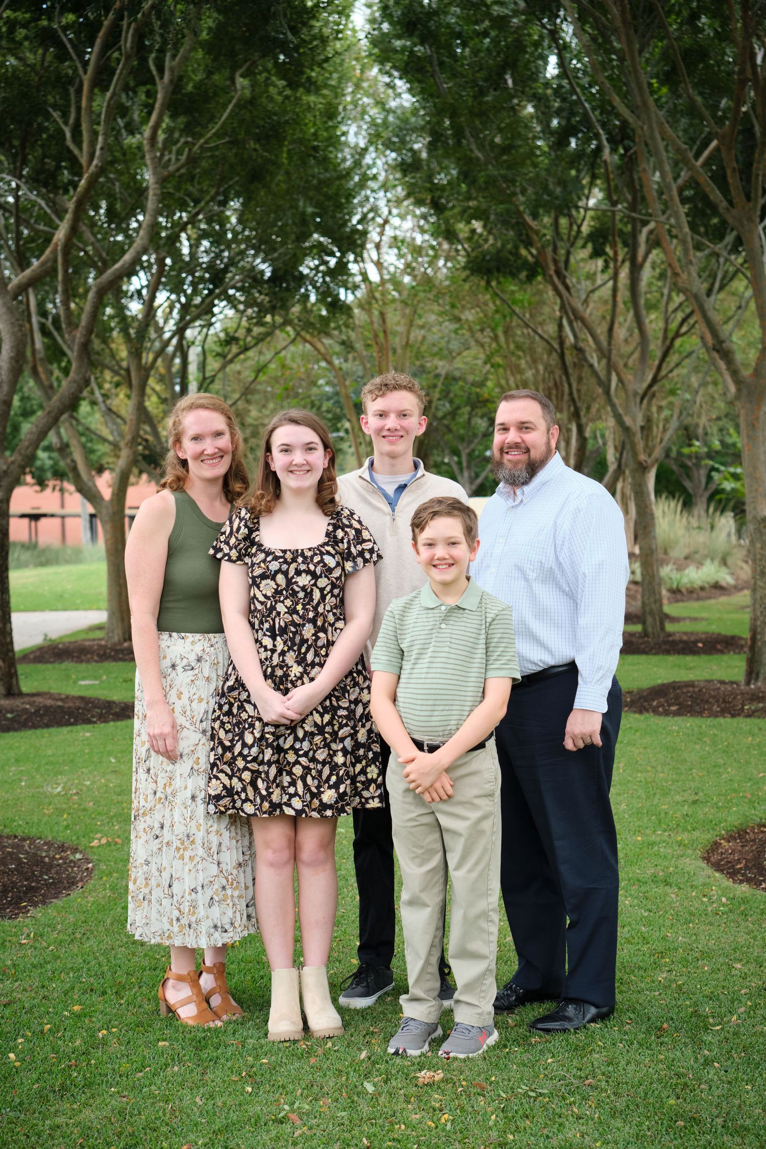 A smiling family of five poses outdoors on green grass surrounded by trees during a warm-weather family portrait session.