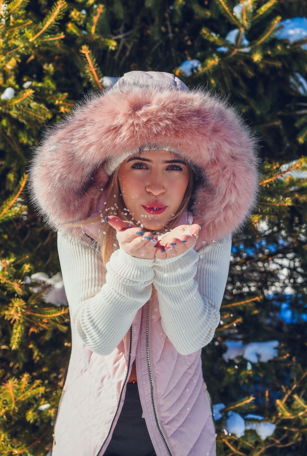 Woman in pink fur hood coat blowing glitter from hands outdoors in snowy winter setting near evergreen tree.