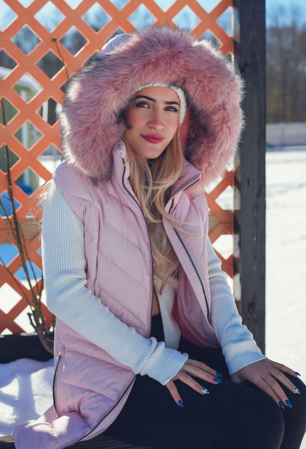 Young woman in pink fur-hooded puffer vest sitting outdoors in snowy winter setting, wearing pink lipstick.