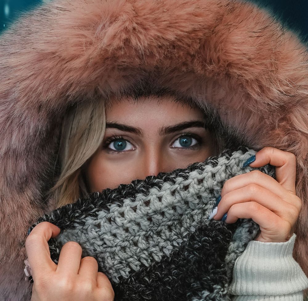 Woman with blue eyes wearing a cozy fur-trimmed hood and gray knit scarf pulled over her face in winter.