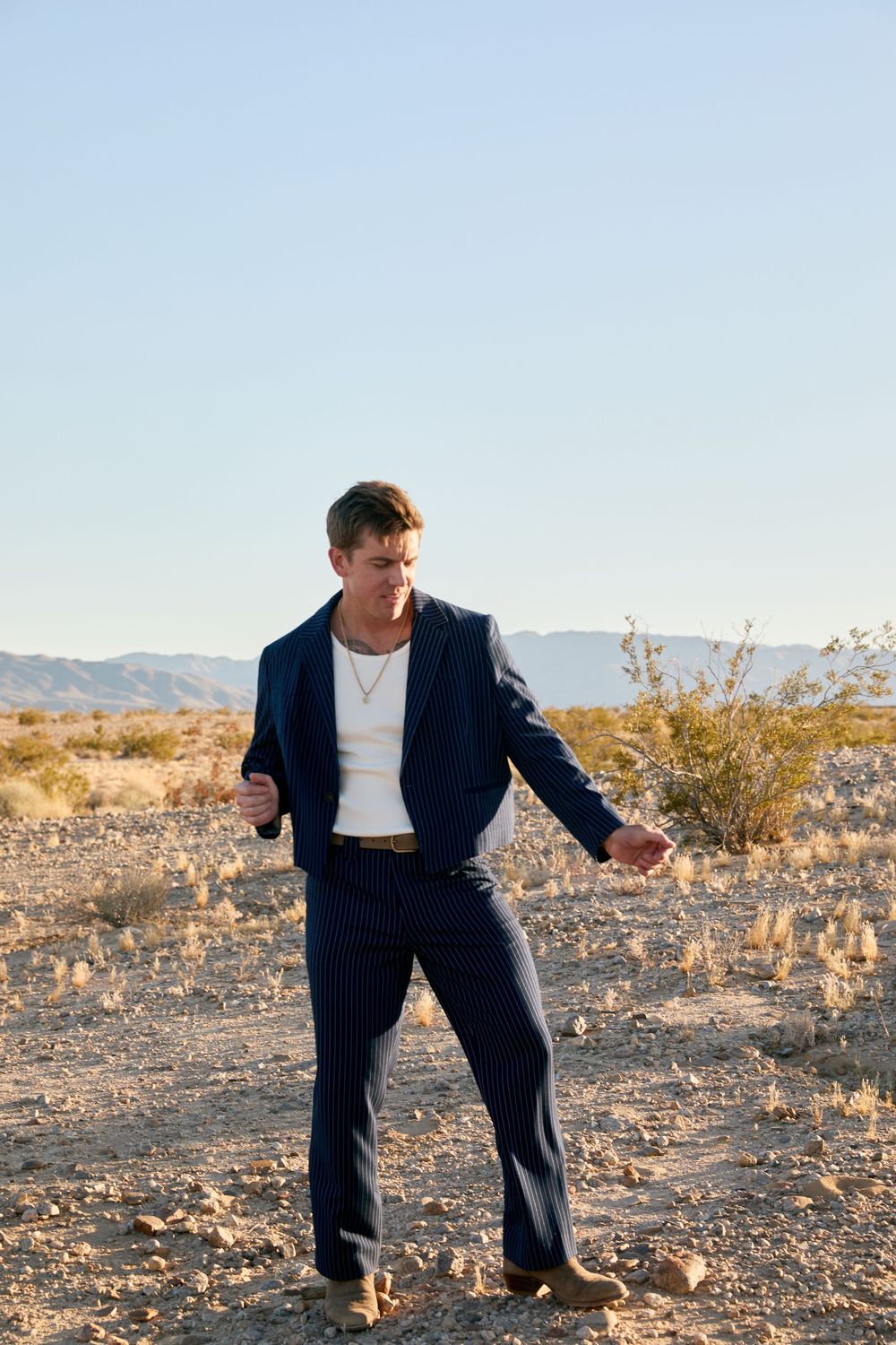 Man in navy pinstripe suit and white tee poses in desert landscape with mountains in background.
