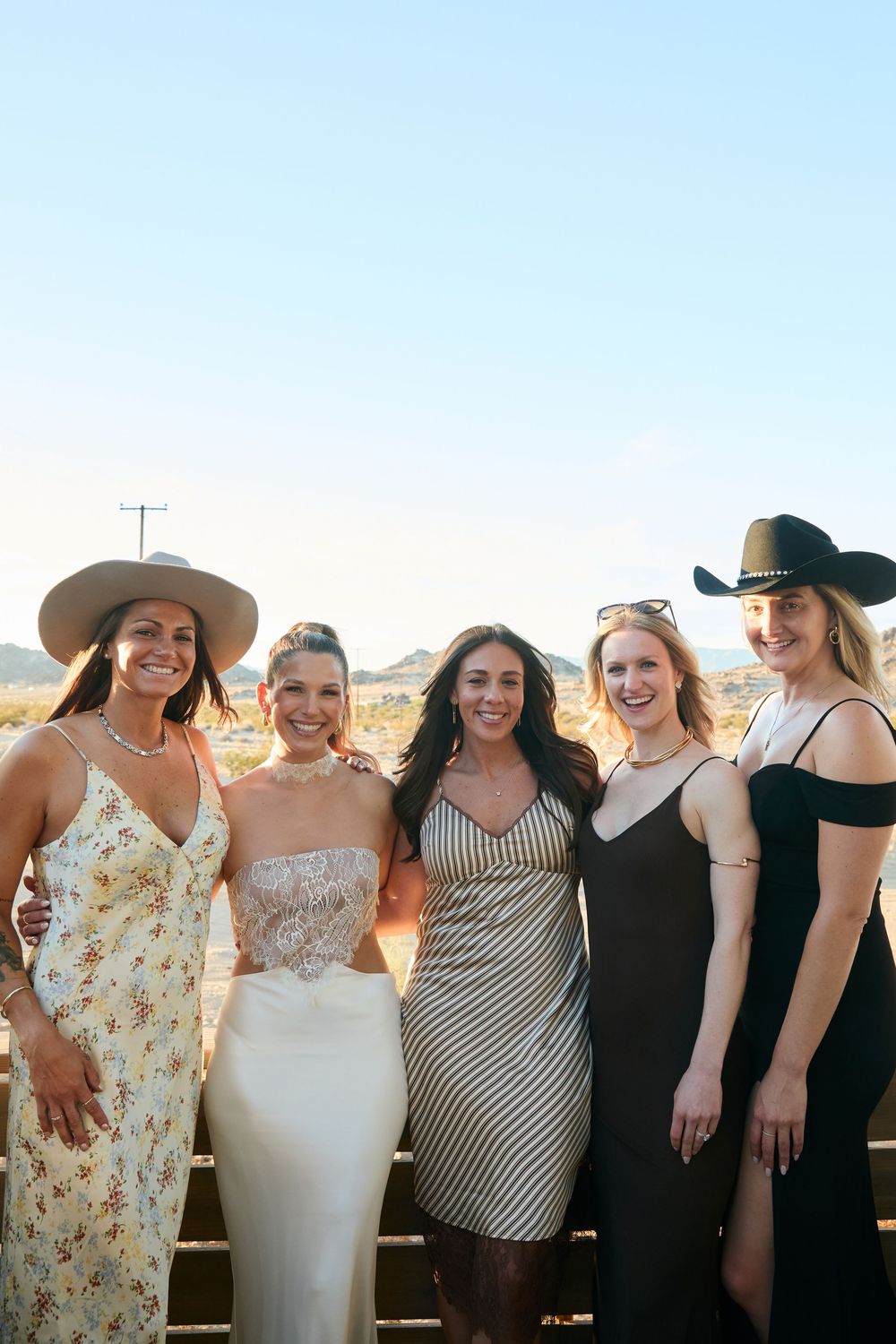 Five women in summer dresses and cowboy hats smiling together outdoors in a desert landscape at golden hour.