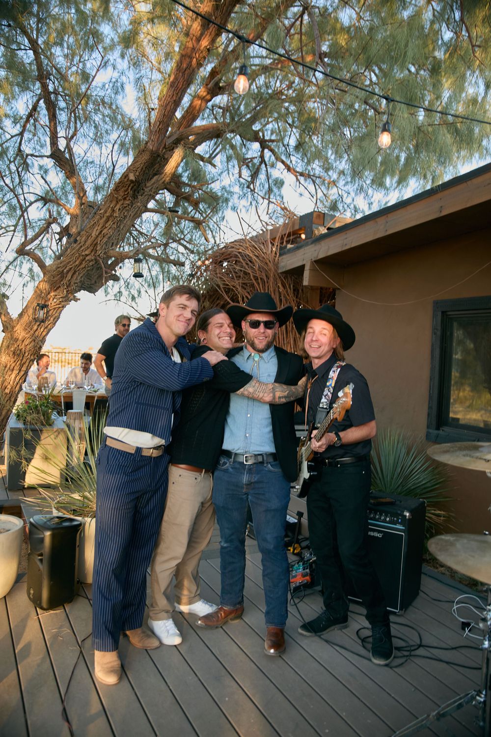 Four men pose together smiling on a wooden deck at an outdoor party with string lights and a guitarist nearby.