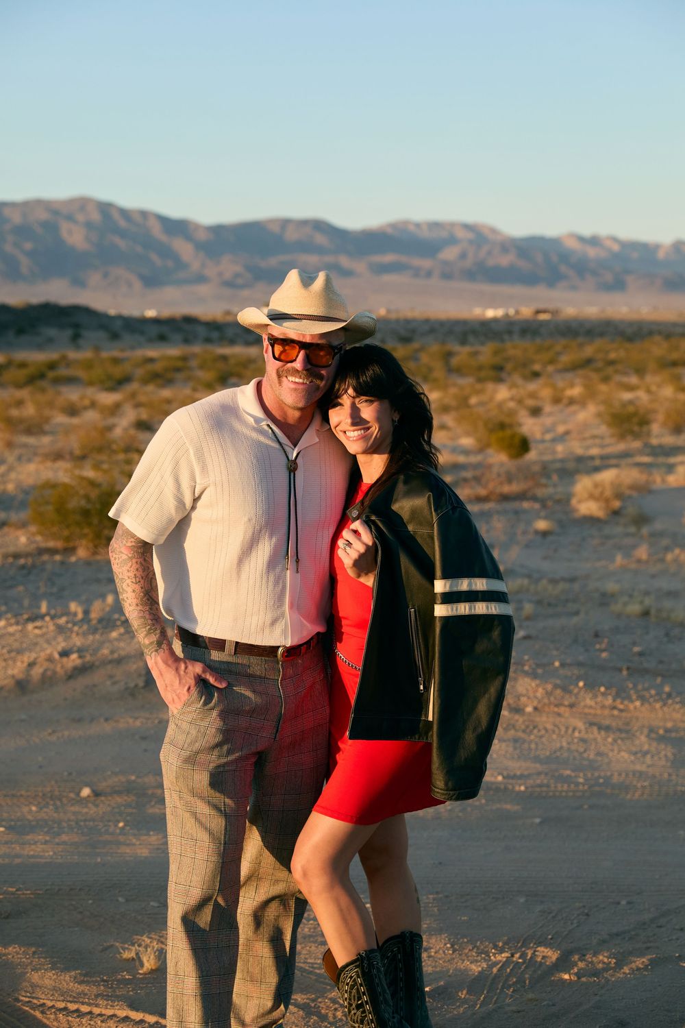 Couple posing in desert landscape at golden hour, man in cowboy hat and woman in red dress with mountains behind.