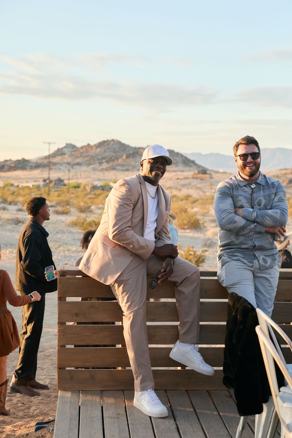 Two men smile and pose on a wooden deck in a desert landscape at golden hour, one in a beige suit, one in gray.