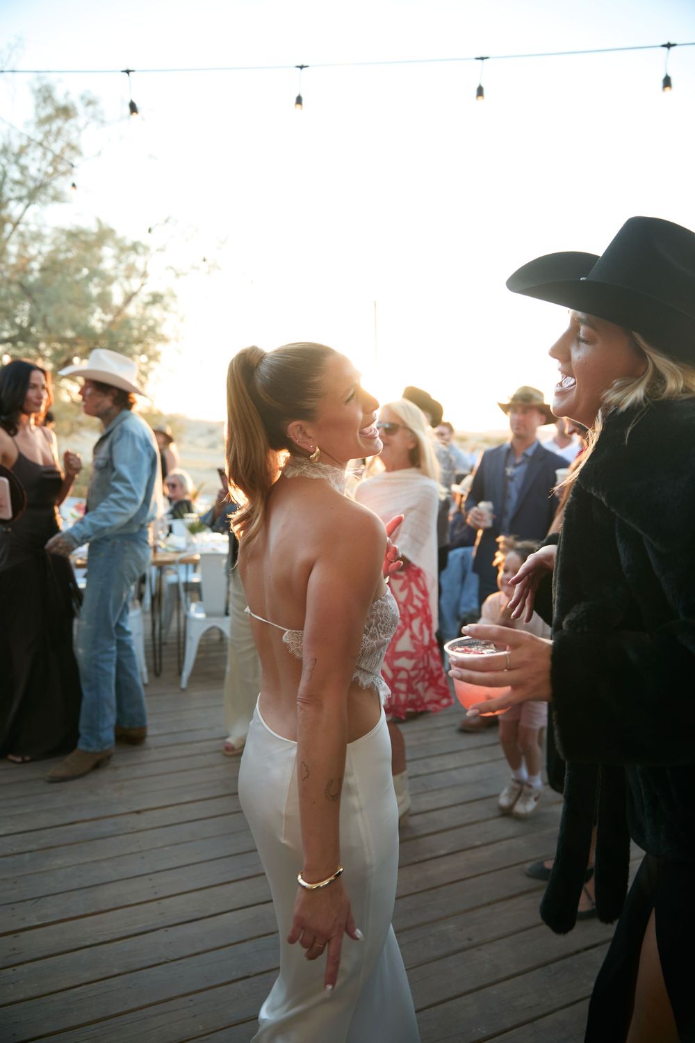 Bride in white gown laughing with guest in black cowboy hat at outdoor wedding reception at golden hour.