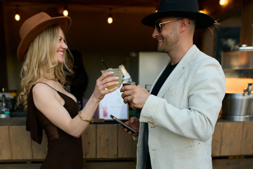 A man and woman in stylish hats toasting drinks together at an outdoor bar venue.